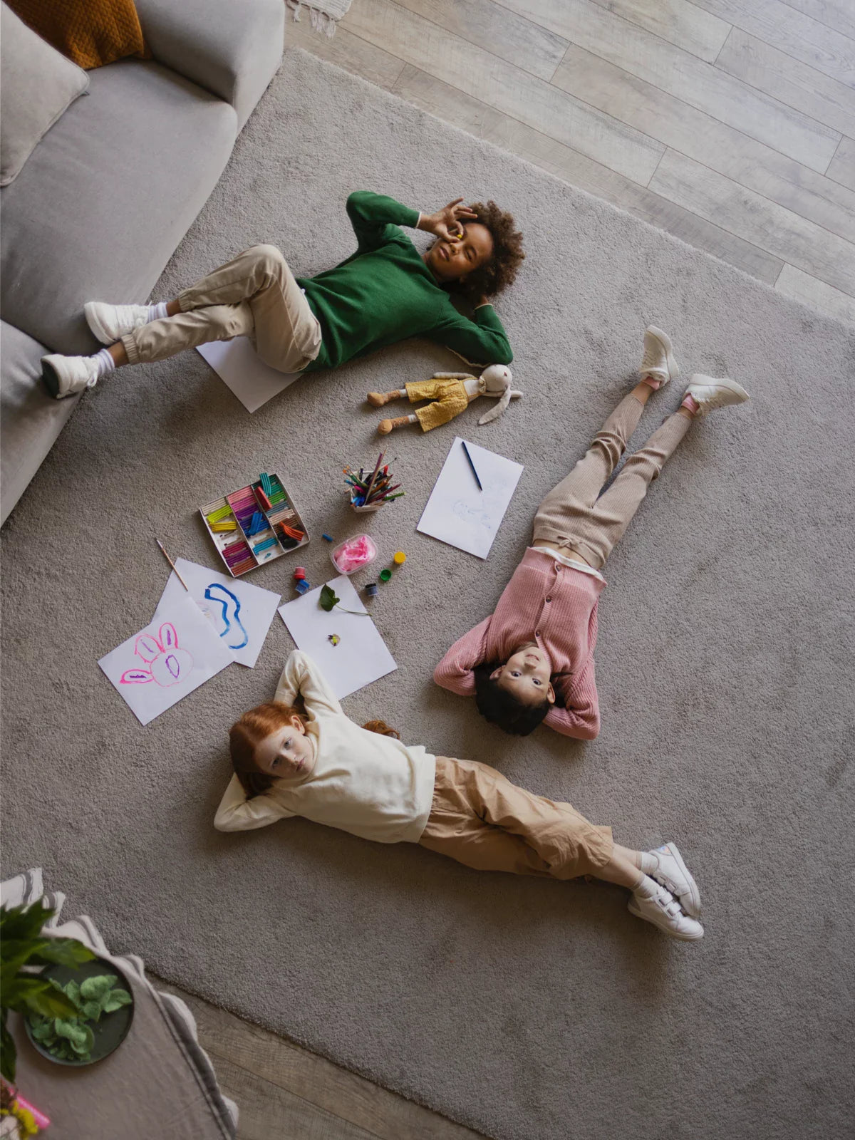Three children lying on a soft, neutral-toned area rug, surrounded by art supplies and drawings, creating a cozy and playful living space.