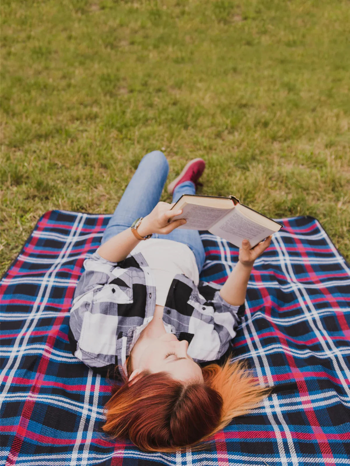 Young woman reading a book while lying on a blue and red plaid outdoor rug in a grassy field, showcasing a comfortable and stylish outdoor living experience.