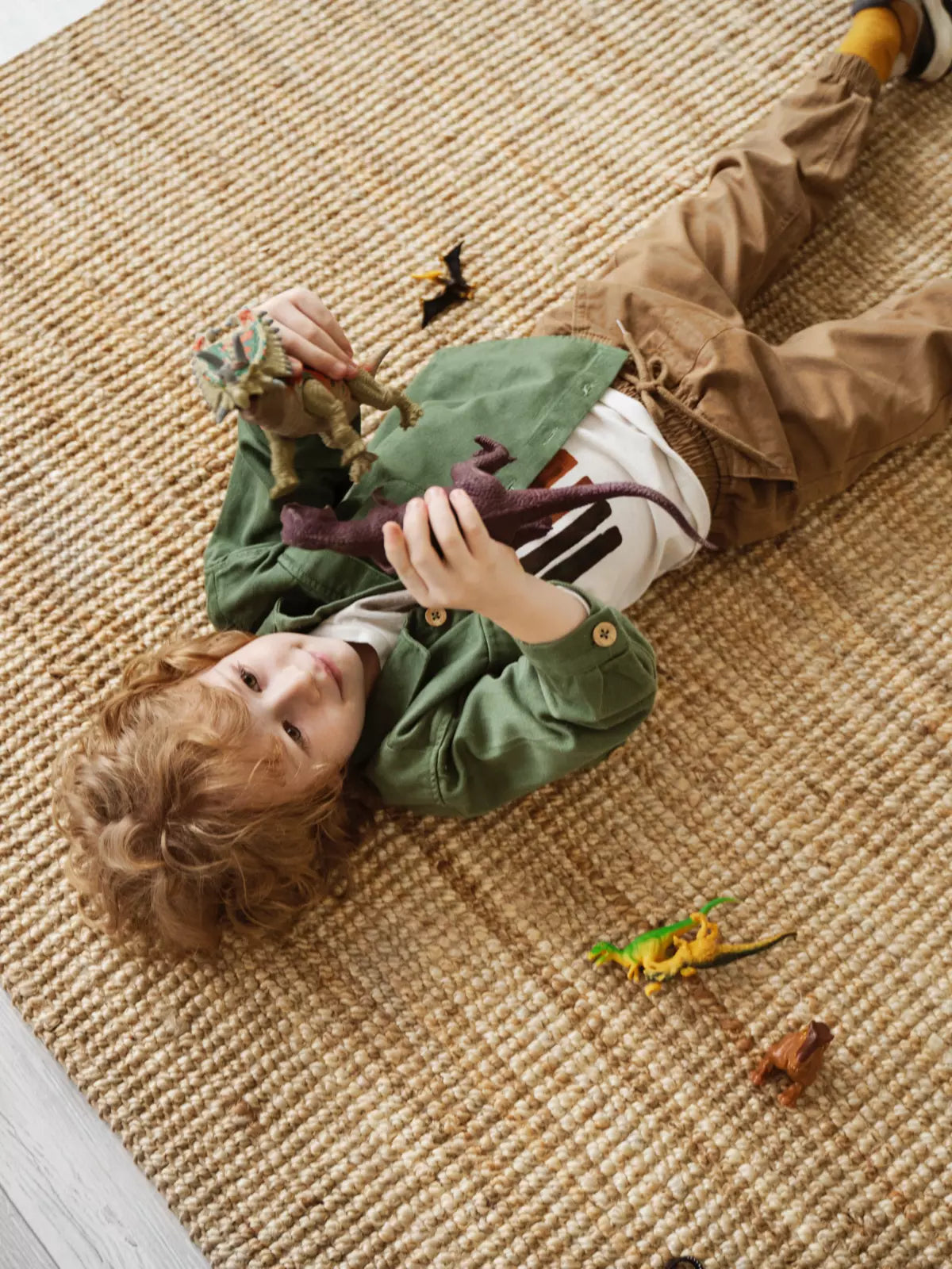 Young child lying on a textured, washable rug, playing with colorful dinosaur toys in a cozy and playful kids’ room setting.