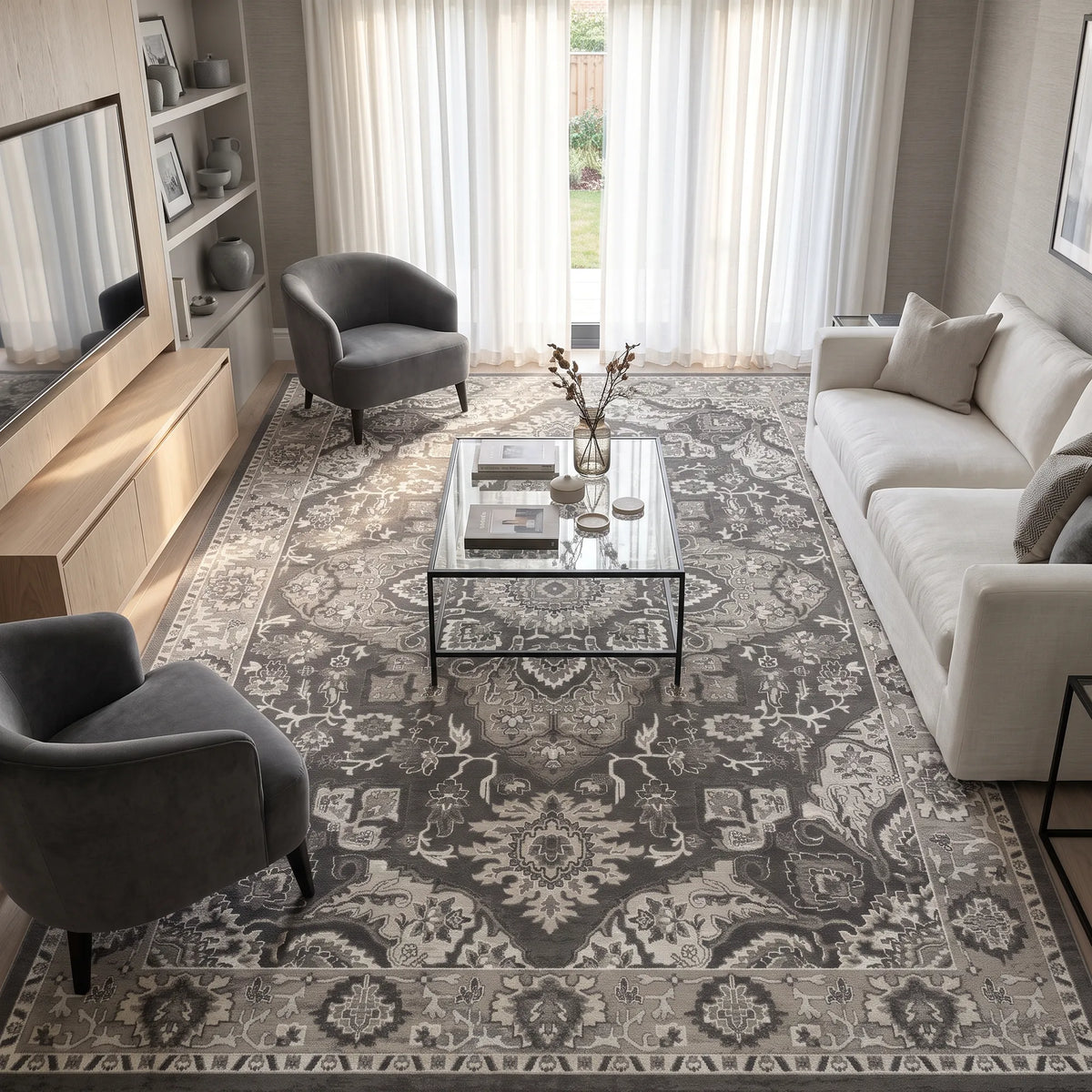 A large traditional-style area rug in gray, and off-white tones, styled in a modern living room with a glass-top coffee table, a light gray sofa, two gray armchairs, and a wooden media console.