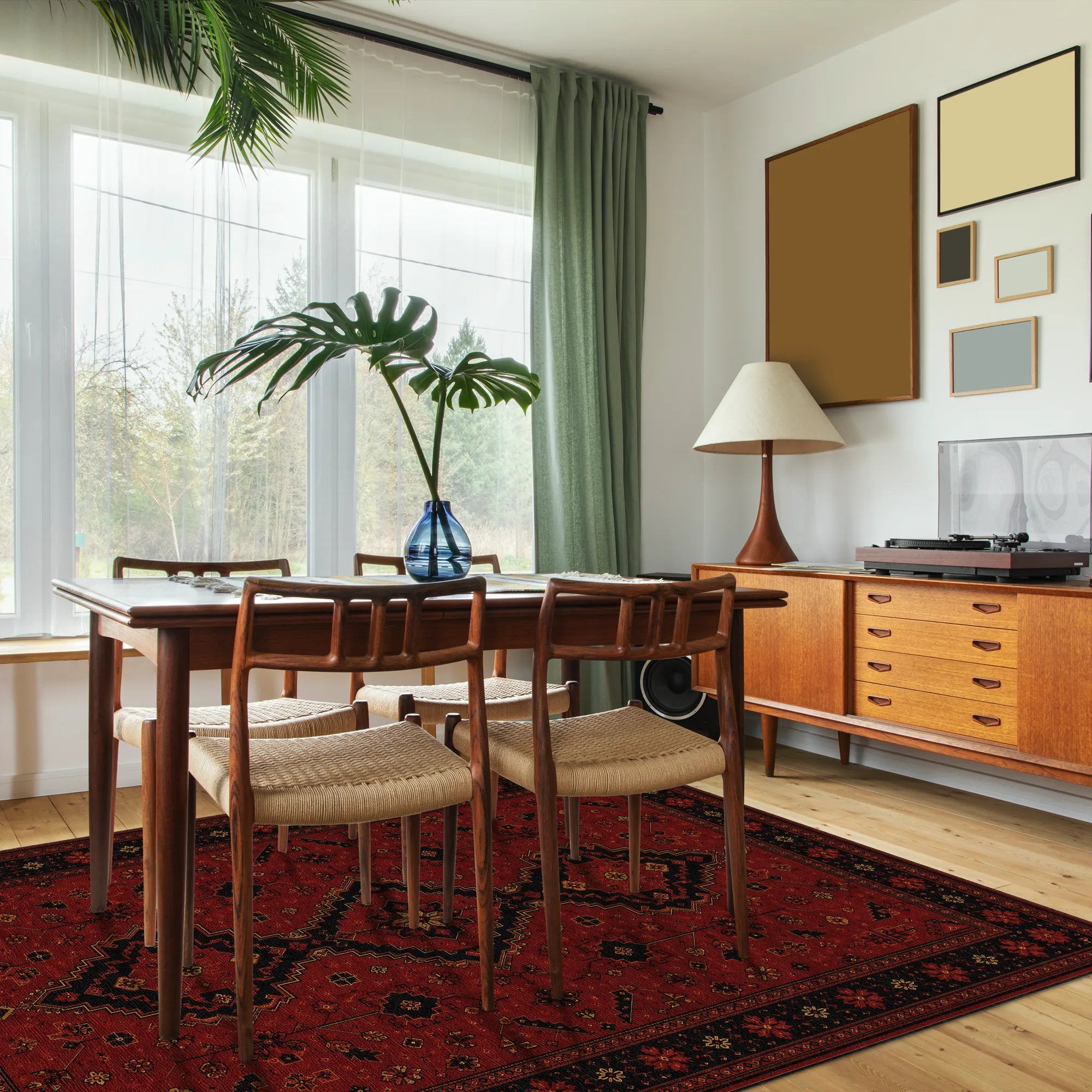 A mid-century modern dining room featuring a wooden dining table and four woven-back chairs on a red patterned rug. The room has light wood flooring, a wooden sideboard with a lamp and record player, and a large window with green curtains.